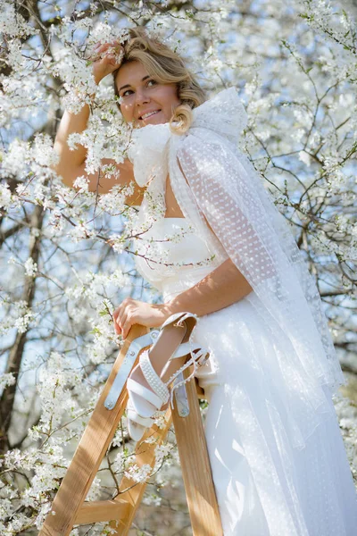 迷人的女士穿着婚纱 肩上戴着大蝴蝶结 站在白色樱花间的木制台阶上 微笑着 一双用珍珠装饰的新娘高跟鞋挂在梯子上 垂直方向 — 图库照片