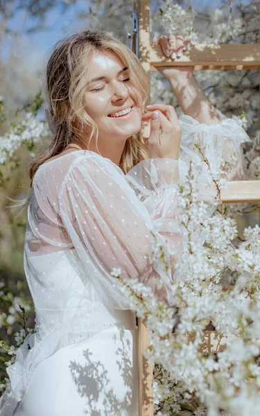 穿着婚纱的年轻貌美的女人站在木制长椅上 紧闭双眼 欣赏大自然 金发碧眼的新娘 周围环绕着洁白的樱花 温柔地微笑着 垂直方向 — 图库照片