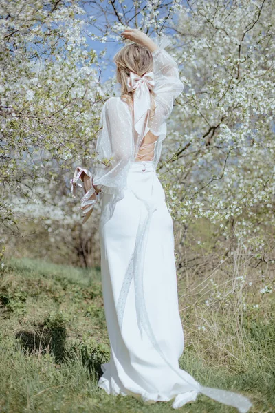 バック桜の木の近くを歩いて上の文字列と結婚式のドレスで若い女性のリアビュー 髪に弓とブロンドの花嫁 ハイヒールの靴のペアを保持し 頭の上に手を持ち上げる — ストック写真