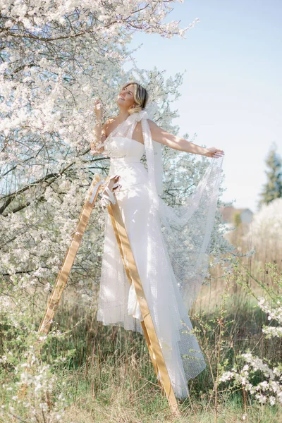 快乐的金发女人穿着华丽的婚纱 高高地站在木制台阶上 望着樱花洁白的花朵 年轻女士手里的衣服是半透明的 垂直方向 — 图库照片