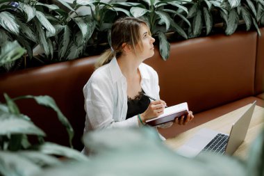 Beyaz gömlekli, siyah dantelli bir elbise giymiş düşünceli bir kadın. Düşüncelere dalmış, günlüğüne notlar yazarken gözlerini kaçırıyor. Kanepede dizüstü bilgisayarın önünde oturan serbest çalışan biri. Bitkilerle çevrili..