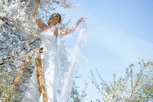 穿着婚纱可爱的金发女士站在白色樱花下的木制台阶上 微笑着望着远方 新娘手里拿着轻薄的婚纱 一双新娘的鞋子挂在梯子上 — 图库照片