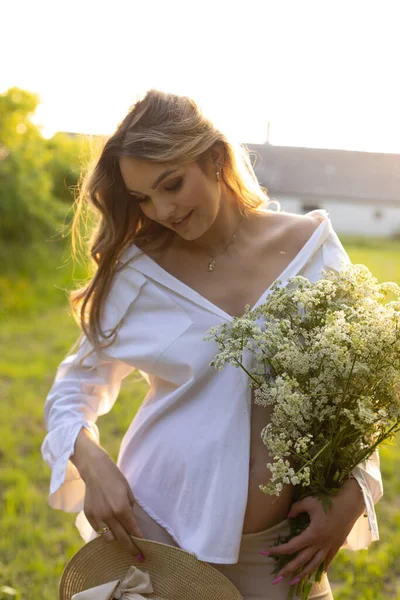 美丽的怀孕少女 留着蓬松的长发 穿着白衬衫 抱着野花 享受着日落的自然 怀孕和做母亲 在等孩子幸福与宁静 — 图库照片