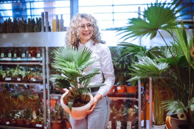 Kotlu ve kazaklı, gülümseyen sarışın elinde palmiye kabıyla bahçe merkezinde duruyor. Kış bahçesi için gösterişsiz bitkiler. Hobi olarak. Tesislerin başarılı alımı. 