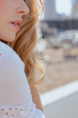 Güneşli bir günde, yüksek binaların çatısında yürürken, sarı saçlı, beyaz elbiseli genç bir kadının yüzünü kapatın. Dalgalı bukle bukleli bir kadın omzunun üstünden bakıyor. Dikey.