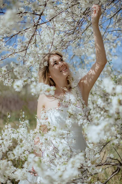 美丽的新娘穿着婚纱 在阳光灿烂的日子走在白樱花旁 金发碧眼的年轻女子被盛开的樱花枝头环抱着 开心地笑着 垂直方向 — 图库照片