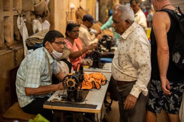 Goa, Hindistan, Şubat 2023. Orta yaşlı, tek kullanımlık maske takan Hintli bir adamın yan görünüşü dikiş makinesinde oturup, kıdemlilerle konuşması kalabalık, geleneksel iş yerlerinde sipariş vermesi..