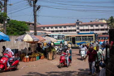 Goa, Hindistan, Şubat 2023. Farklı insanlarla dolu kalabalık bir yol, otobüs durağı yakınında motosikletliler, büyük şemsiyelerin altında sebzeli tezgahlar, güneşli bir günde yerel geleneksel pazar.