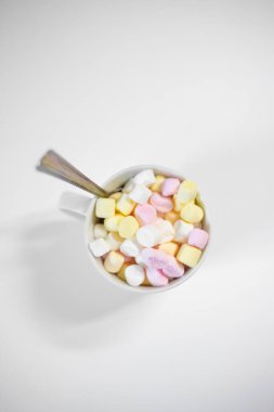 Bardağın yukarısından çok renkli küçük şekerlemeler ve çay kaşığı beyaz boş masada duran çay kaşığının görüntüsü. Beyazların arka planında yumuşak renkli şekerlerle dolu beyaz bir kase. Dikey.