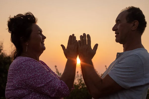 Wonderful mature couple standing facing each other against sunset, folding palms together and joining hands, practicing yoga pose in nature. Woman happily looking at calm bald man in front.