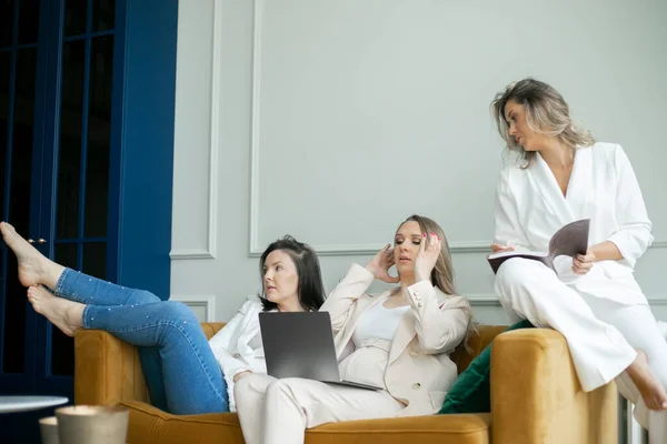 Three business ladies sitting on yellow sofa and working together. Pregnant woman with laptop on lap straightening hair while colleague with journal looking at monitor, brunette distracted from work.