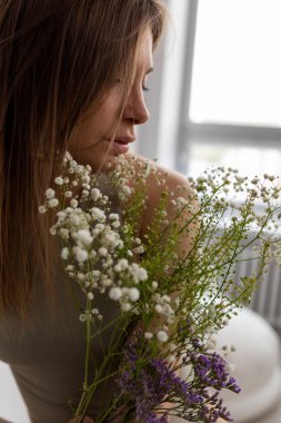 Uzun siyah saçlı güzel bir kadının yan görünüşü bej mayo giyer, pencerenin yanında durur, elinde yeşil bitki çoban çantası, lavanta ve çingene buketi taşır. Dikey, yumuşak odaklanma. Doğa.