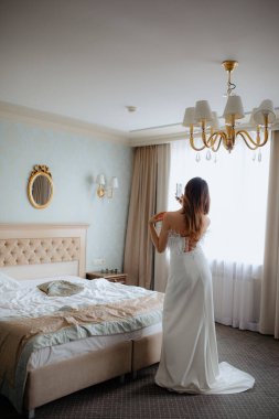 Uzun gelinlikli esmer, çıplak omuzlu, otel odasındaki geniş çift kişilik yatağın yanında elinde telefonla duruyor. Büyüleyici gelin aynada asılı duruyor..