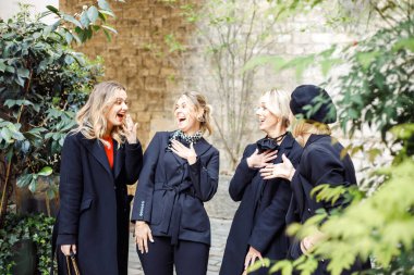 Siyah elbiseli, açık renk elbiseli, yeşil ağaçların yanında dikilen, beklenmedik dedikodulardan şok olmuş genç ve harika kadın arkadaşlarının portresi..