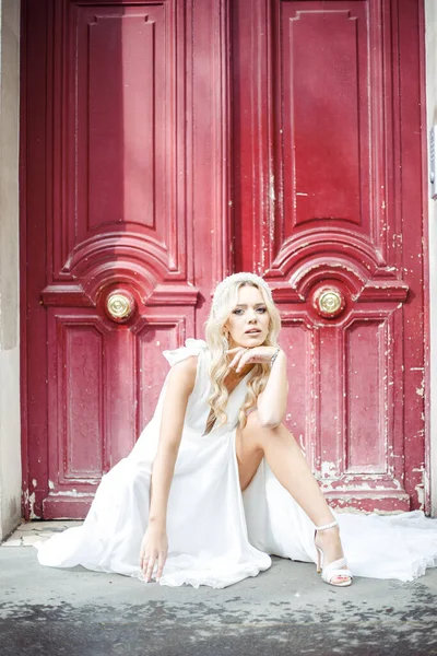 Portrait of young wonderful woman with fair hair wearing long white light dress, decorated hairband, sitting on street near old red wooden doors, leaning hand on bare knee. Beauty, wedding. Vertical.