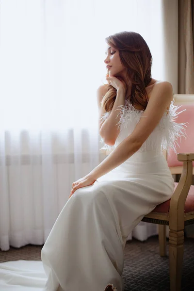 Elegant young woman with dark hair wearing long shoulderless wedding dress decorated with little feathers sitting on chair next to curtains. Lady softly touching face while looking aside.