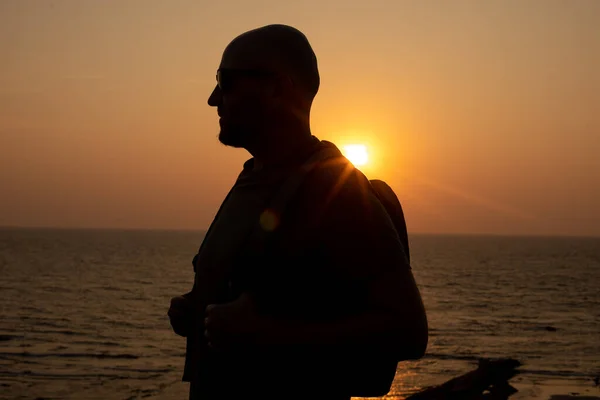 Portrait of bald man in sunglasses walking with bag on back near calm sea on seashore at sunset. Traveler with backpack on shoulders standing against seascape at end of summer day.