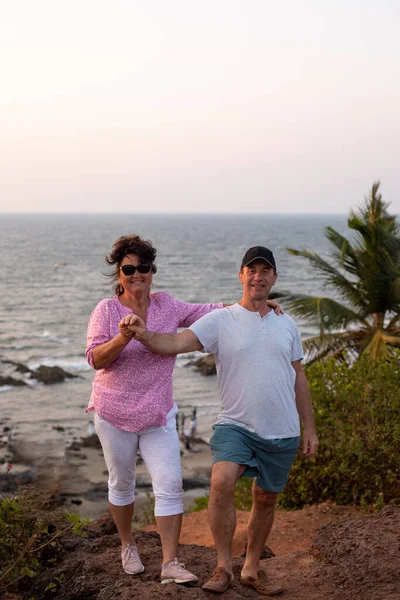 Delightful couple standing on hill against seascape. Smiling lady in pink t-shirt and sunglasses holding male hand and keeping arm on shoulder of joyful man in black cap and white t-shirt. Vertical.