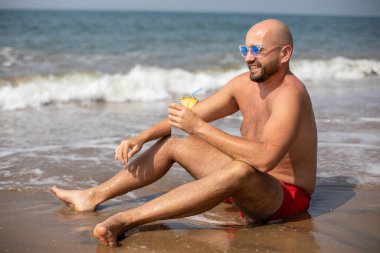Mavi güneş gözlüklü genç tatilci dalgalı denizin yanındaki kumlu sahilde dinleniyor ve gülümsüyor. Kırmızı mayo giymiş sakallı üstsüz bir adam elinde ananas parçasıyla süslenmiş bir içecek tutuyor..