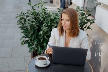 Çekici, kahverengi saçlı, zarif ceketli serbest çalışan bayan açık hava kafedeki terasta kahve içer ve dizüstü bilgisayarda çalışır. Başarı ve kariyer. Kendi işin ve girişimciliğin.. 