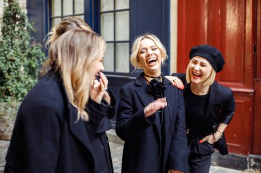 Siyah elbiseli, açık renk elbiseli, dışarıda, renkli duvarların yanında duran, etkileşimli kıkırdayan, komik dükkanlar paylaşan genç ve güzel kadın arkadaşlarının portresi..