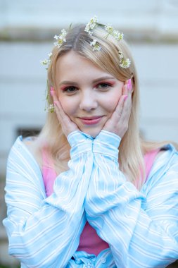 Saçları beyaz çiçeklerle süslenmiş, mavi gömlekli, avuçlarını yüzüne bastıran güzel sarışın kızın yakın plan portresi. Gençlik ve güzellik. Pozitif duygular. 