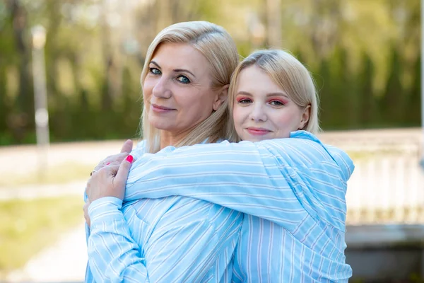 Daughter hugs mom in park for walk. They are wearing identical blue cotton shirts. Positive emotions. Family relations. Closeness and understanding. Love and trust. Connection of generations.