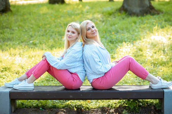 Two blonde mom and daughter dressed in same clothes are sitting on wooden bench in park with their backs pressed against each other and looking into camera. Family relations. Symmetrical image.
