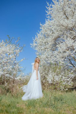 Güneşli bir günde, beyaz kiraz çiçeklerinin arasında uzun bir gelinlikle sırtı iplerle gezen lüks genç bir kadın. Güzel bir gelin, sarı saçlı, arkası da harika bir gülümsemeyle bakıyor. Dikey.