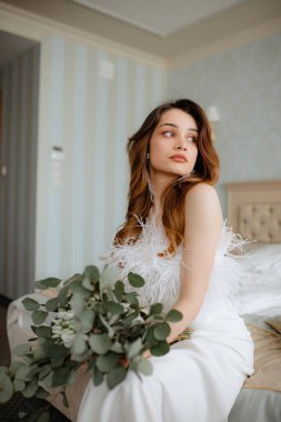 Kırmızımsı kahverengi dalgalı saçlı, gelinlik giymiş tüylerle süslenmiş, elinde çiçeklerle yatakta oturan genç bir bayan. Tassel küpeli hoş gelin başka tarafa bakıyor..