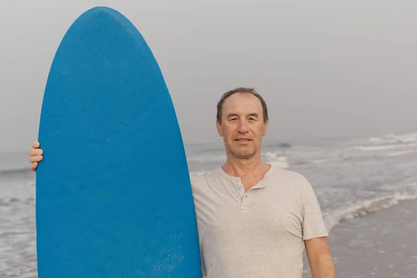 Portrait of sporty middle aged surfer wearing light gray polo shirt standing on waving seaside against seascape and holding blue surfboard close to body. Sportsman with surfing board looking ahead.