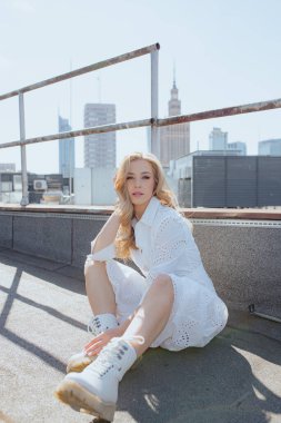 Nakış işlemeli beyaz pamuklu elbise giyen çekici bir kadın ve yüksek binaların çatısında çitin önünde oturan bağcıklı botlar. Sarışın, şehrin arka planında baş aşağı duruyor. Dikey.