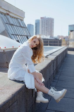 Sarışın, beyaz elbiseli hoş bir kadın yüksek binaların çatısında sallanan bacaklarıyla oturuyor. Pamuklu elbiseli ve beyaz çizmeli genç bayan evin tepesinde dinleniyor. Dikey.
