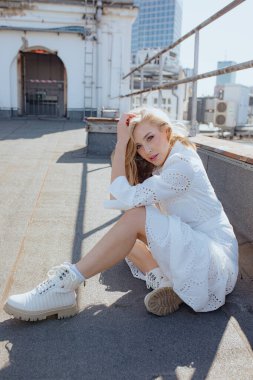 Beyaz pamuklu gecelikli, sarı dalgalı saçlı, işlemeli sevimli genç bayan, yüksek binaların güneşli çatısında oturan ve kilitlere elle dokunan bağcıklı botlar. Dikey.