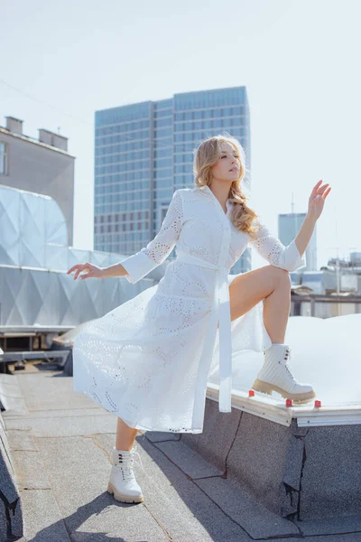 Rhythms ofcity. Urban fashion. Flirtatious blonde in white dress poses, showing off her leg in stylish white shoes, on roof of building against backdrop of urban landscape. Modern free women.