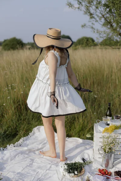 stock image Unrecognizable young girl in snow-white summer dress and hat enjoys summer day in nature, stands barefoot on blanket among tall green grass. Freedom, relaxation. Bleachers, conditioners for laundry.