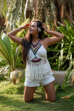 Beyaz egzotik çiçeklerle süslenmiş uzun siyah saçlı, beyaz bluzlu, şortlu, palmiye ağaçlarının yanındaki yeşil çimlerin üzerinde duran genç ve mutlu Asyalı kadının portresi. Güzellik, seyahat.