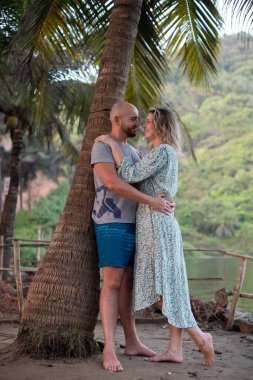 Çıplak ayakla ayakta duran genç bir çiftin portresi, nehir kenarında kucaklaşan, yeşil dağlar. Kel adam gülümseyen kadına sarılıyor, palmiye ağacına yaslanıyor, tatilin tadını çıkarıyor, gözlerle buluşuyor. Seyahat, güzellik.