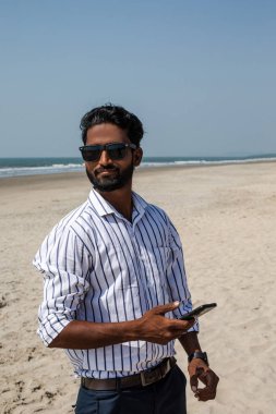 Çizgili gömlek giyen, siyah güneş gözlüğü takan, deniz kıyısındaki kumsalda elinde akıllı telefon tutan genç Hintli bir iş adamının portresi. Seyahat, egzotik tatil beldesi, turizm, yaz.