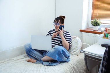 Okuma gözlüklü ve rahat ev kıyafetleri giyen hoş bir kadın evde dizüstü bilgisayarla çalışırken cep telefonuyla konuşuyor. İş kadını. İnternette çalışıyorum. Yaşam biçimi. 