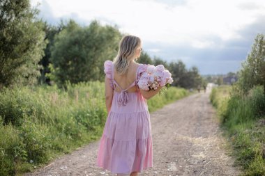 Yazın kır yolunda, elinde bir buket soluk pembe şakayık demeti olan tanınmayan bir kadının görüntüsü. Kadınlık ve güzellik. Olumlu duygular. Sakin bir ruh hali. Çiçek yetiştirme işi. 