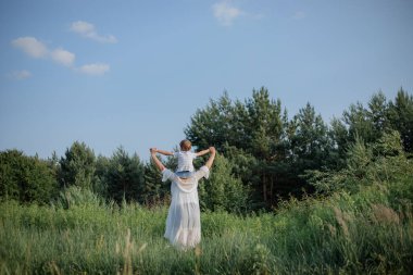 Anne, yaz kıyafetleri içinde, küçük oğlunu omuzlarında tutarken, yaz günü tarlada yürür. Bekar ebeveynler. Aile ilişkileri. Birlikte mutluyuz. Köyde tatiller.