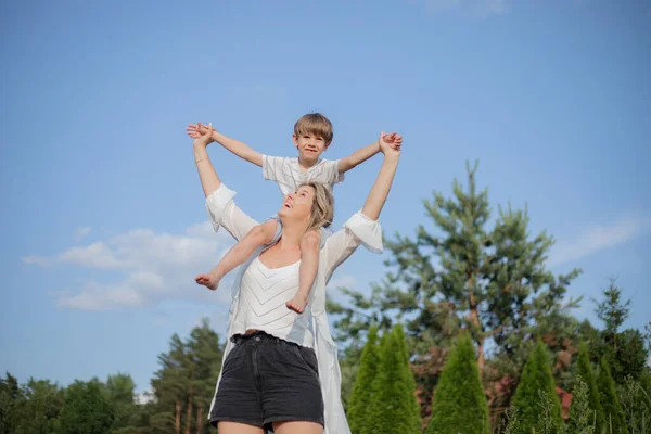 Positive emotions. Happy mother in summer clothes holds her little son on her shoulders, he enjoys height. Single parents. Family relations. Happy together. Love, understanding. Holidays in village.
