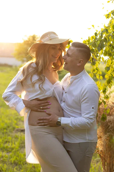 Young man in white shirt laughs happily and hugs his pregnant beautiful wife by nature in rays of setting sun. Positive emotions. Wonderful moments. Family happiness. Waiting for child.