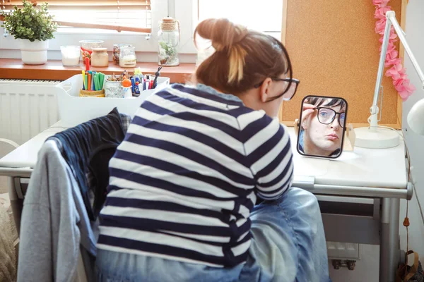 Young woman in home clothes is sitting with her back to camera looking into desk mirror, to which her face is reflected. Home life. Beauty treatments at home. Home interior. Lifestyle.