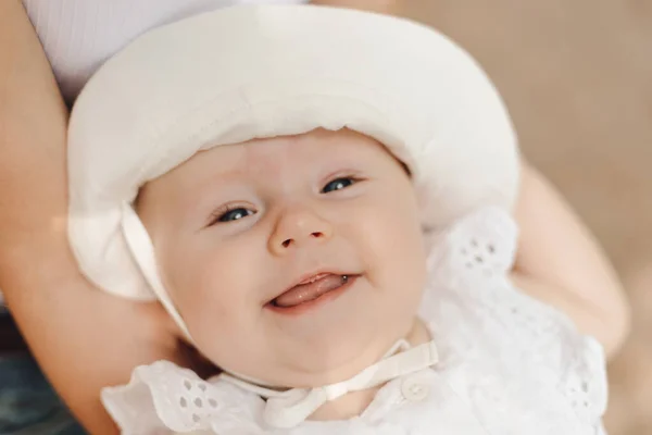 Close-up portrait of smiling toothless newborn girl in white panama hat, held in arms of unrecognizable mother. Newborn care. Hygiene products for children. Nutrition of newborns. Medical insurance.
