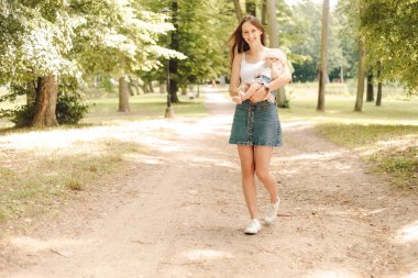 Yeni doğmuş bir bebekle yürüyorum. Kısa kot etekli ve kollarında küçük kızı olan beyaz tişörtlü genç anne yaz gününün ve keyfinin tadını çıkarıyor. Bekar anne. Yeni doğan bakımı. Sağlık sigortası .