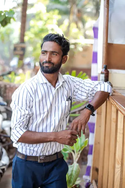 Portrait of confident young well-groomed Indian man businessman wearing striped shirt, blue trousers, leaning on bar counter, looking aside, smiling, enjoying weather. Travelling, exotic resort.