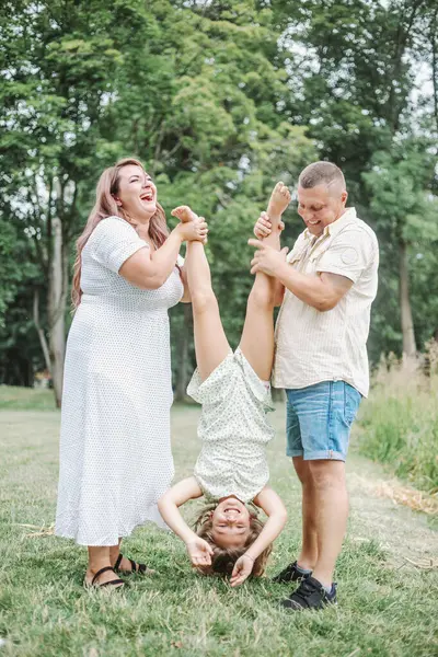 Happy childhood. Full-length vertical portrait, mom and dad are standing in nature, laughing and holding their daughter by legs, who is hanging upside down.Harmonious loving happy family. Love trust.