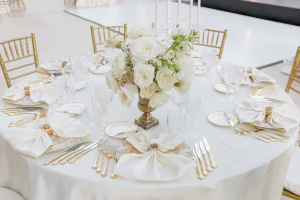 Round table at wedding event for six people with classic dishes with gold drawings, gilded silverware, white table textiles, crystal and bouquets of roses. Organization, decoration of celebrations.